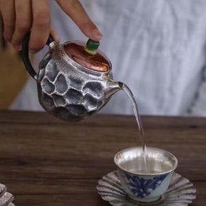 Puede incluir: Una tetera plateada con un dise&ntilde;o texturizado en forma de panal vierte agua en una taza de t&eacute; a juego. La tetera tiene un asa negra y una tapa de color cobre con un pomo verde. La taza y el platillo tienen un estampado floral azul.