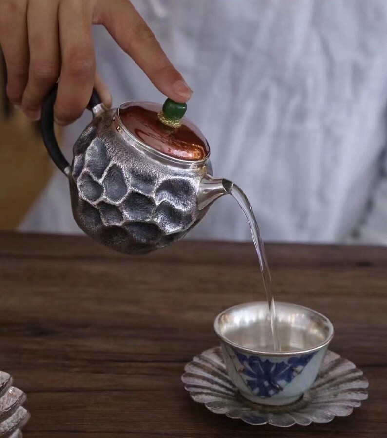 Puede incluir: Una tetera plateada con un dise&ntilde;o texturizado en forma de panal vierte agua en una taza de t&eacute; a juego. La tetera tiene un asa negra y una tapa de color cobre con un pomo verde. La taza y el platillo tienen un estampado floral azul.