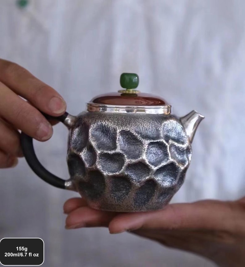 Puede incluir: Tetera plateada con un patr&oacute;n texturizado en forma de panal. Tiene un asa oscura, una tapa marr&oacute;n y una piedra verde en la parte superior. La tetera tiene una capacidad de 200 ml y pesa 155 g.