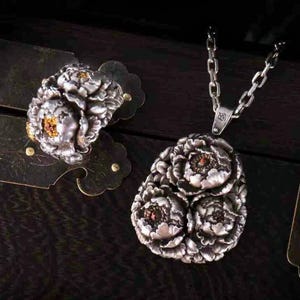 Puede incluir: Un conjunto de anillo y collar con colgante de plata. El anillo y el colgante presentan diseños florales detallados con pequeños acentos de color naranja. El collar tiene una cadena de plata. Las joyas se exhiben sobre una superficie de madera oscura.