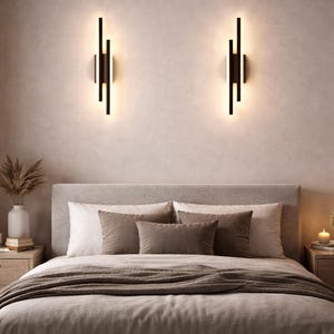 Op de afbeelding: Slaapkamer met een bed, kussens en een bruin deken. Twee moderne zwarte wandkandelaars met warme verlichting zijn boven het bed gemonteerd. De kamer heeft een neutraal kleurenpalet.