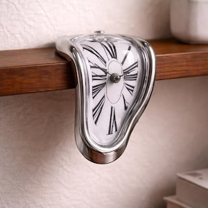 Puede incluir: Un reloj plateado y derritiéndose con una esfera blanca y números romanos negros. El reloj de estilo surrealista se exhibe en una estantería de madera. Una pequeña planta en maceta y libros están en el fondo.