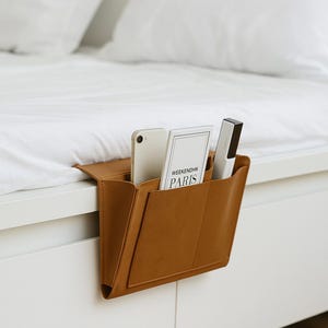 May include: A tan leather bedside organizer hanging from a white bed frame. The organizer holds a smartphone, a book with "WEEKEND IN PARIS" text, and a remote control. The bed has white linens.