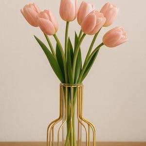 Könnte beinhalten: Ein Strauß zartrosa Tulpen in einer goldfarbenen Drahtvase. Die Vase hat einen transparenten Glaseinsatz. Die Tulpen haben grüne Stiele und Blätter. Der Hintergrund ist in einem neutralen Beige gehalten.