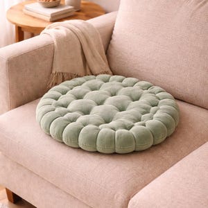 Könnte beinhalten: Ein runder, salbeigrüner, getufteter Kissenbezug liegt auf einem hellrosa Sofa. Das Kissen hat eine weiche, samtige Textur und ein knopfartiges Design. Das Sofa ist Teil einer Wohnzimmereinrichtung, mit einem Beistelltisch und einer Decke.