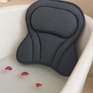 Può includere: Un cuscino da bagno grigio scuro è posizionato in una vasca da bagno bianca piena d'acqua. Il cuscino ha una superficie strutturata e una forma sagomata per il supporto di collo e schiena. Petali di rosa rossi galleggiano nell'acqua, creando un'atmosfera rilassante.