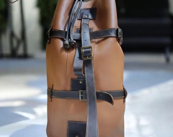 Könnte beinhalten: Ein brauner Lederrucksack mit schwarzen Riemen und Metallschnallen. Die Tasche hat einen Kordelzugverschluss an der Oberseite und einen Griff. Das Design umfasst mehrere Riemen und eine kleine quadratische Tasche. Die Tasche ist aus Leder.