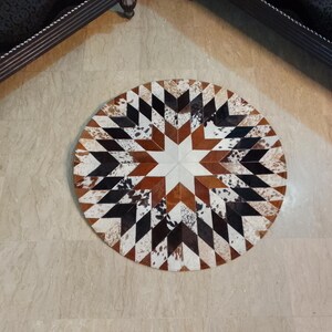 Può includere: Tappeto rotondo in pelle di mucca con un motivo geometrico a stella. Il tappeto presenta una stella bianca centrale circondata da triangoli marroni, neri e bianchi. Il tappeto ha un diametro di circa 122 cm.
