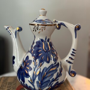 May include: White ceramic teapot with a blue floral design and gold accents. The teapot features a curved handle and spout, and a lid with a gold finial. The floral pattern includes stylized flowers and leaves.