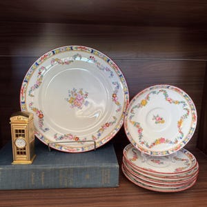 May include: A collection of floral-decorated porcelain tableware, including plates and saucers, with a red trim. A gold-colored miniature telephone booth with a clock sits on a blue book. The plates have a cream background with colorful floral patterns.
