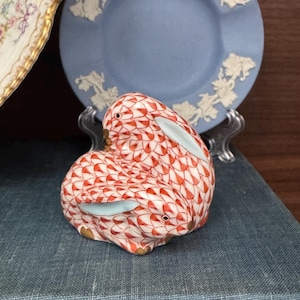 May include: A ceramic figurine of a rabbit, featuring a red and white fishnet pattern. The rabbit has light blue accents on its ears and is posed curled up. A blue plate with white floral detailing is in the background.