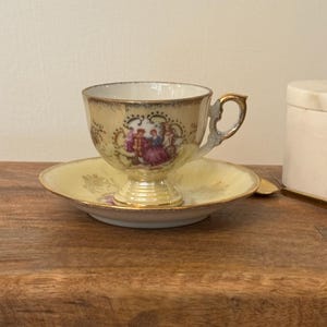 May include: A vintage teacup and saucer set with a yellow base and gold trim. The cup features a floral design with a couple in the center.