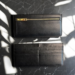 May include: Two black leather wallets are displayed on a marble surface. The top wallet has a gold zipper closure. The bottom wallet is open, revealing multiple card slots. The leather has a textured grain.