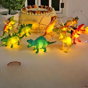 May include: A decorative string of dinosaur-shaped LED lights in green, yellow, and orange. The lights are strung on a thin wire. A cake with white icing and brown decorations is in the background. The lights are suitable for a child's bedroom.