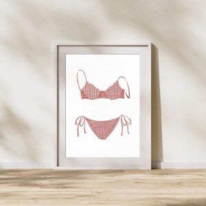 Puede incluir: Impresión enmarcada de un conjunto de bikini a cuadros rojos y blancos. El top del bikini tiene tirantes finos y la parte inferior tiene lazos en los lados. La obra de arte se muestra sobre un fondo blanco.
