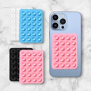 Peut inclure: Plusieurs coussinets à ventouse colorés pour téléphones portables. Les coussinets sont blancs, bleus, noirs et roses. Un coussinet rose est fixé au dos d'un smartphone bleu clair. Les coussinets ont une grille de petites ventouses.