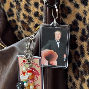 Puede incluir: Llavero acrílico transparente con una foto de un hombre en esmoquin y un segundo llavero con cuentas de colores, dados y un gráfico. Los llaveros están sujetos a un bolso de cuero marrón con un fondo de estampado de leopardo.