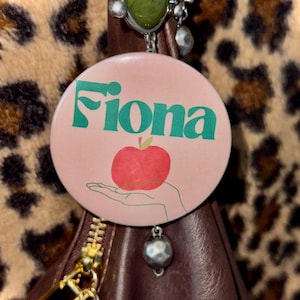 Puede incluir: Un botón redondo rosa con el nombre "Fiona" en verde y una manzana roja. El botón está sujeto a un bolso de cuero marrón con cremallera dorada y un charm decorativo. Un estampado de leopardo es visible en el fondo.