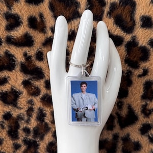 Op de afbeelding: Een heldere, rechthoekige sleutelhanger met een foto van een persoon in een lichtgrijs pak. De sleutelhanger is bevestigd aan een zilveren metalen ring. De achtergrond is een luipaardprint.