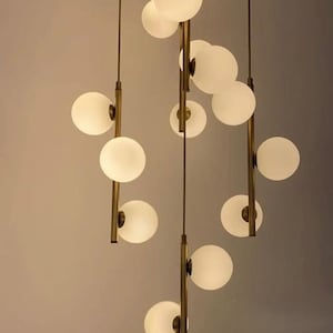 May include: Contemporary chandelier with multiple white globe lights suspended from a brass-coloured canopy. The lights are arranged at varying heights, creating a visually appealing design. Suitable for interior lighting.