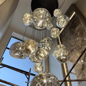 May include: A modern chandelier featuring multiple irregularly shaped, silver-toned glass globes suspended from black cords. The globes reflect light, creating a warm, ambient glow. The fixture is mounted on a black ceiling plate, suitable for interior lighting.