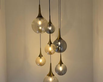 May include: A modern chandelier with six glass globe shades. Each globe has a brass-colored cone-shaped top and hangs from a black cord. The globes are clear and amber-toned, with a light bulb inside.