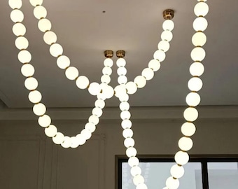 Puede incluir: Una lámpara de araña moderna con múltiples hilos de luces esféricas brillantes. Las luces están dispuestas en un patrón en cascada, suspendidas de soportes de techo de color latón. El diseño es elegante y contemporáneo, adecuado para varios interiores.