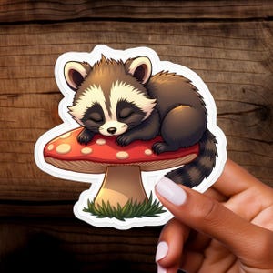 Autocollant Raton laveur endormi sur champignon rouge – Autocollant en vinyle mignon pour ordinateur portable, amoureux de la nature et idées cadeaux