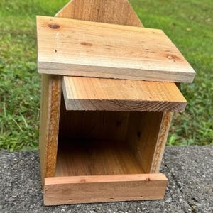 May include: A small, untreated wooden birdhouse with a pitched roof and a rectangular entrance. The birdhouse is constructed from light-coloured wood and is positioned on a grey surface, with green grass visible in the background. Designed to provide shelter for small birds.