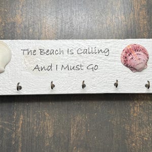 May include: A white wooden key holder with six hooks, decorated with seashells. The text "The Beach Is Calling And I Must Go" is printed on the board. The key holder is approximately 12 inches long.