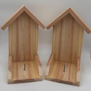 Puede incluir: Dos casas para pájaros de madera con tejados a dos aguas. Las casas para pájaros son de madera de color claro y tienen un diseño simple y rectangular. Cada casa para pájaros tiene una pequeña repisa en la parte inferior y un tornillo visible en el interior.