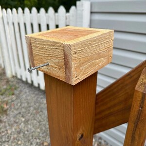 Könnte beinhalten: Ein Holzpfosten mit quadratischem Kopf, befestigt mit einer Schraube. Der Pfosten ist Teil einer Holzkonstruktion, wahrscheinlich ein Deck oder ein Geländer. Das Holz hat ein natürliches, unbehandeltes Aussehen.