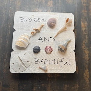 May include: A white wooden sign with the words "Broken AND Beautiful" in black script, adorned with various seashells. The shells include a sand dollar, conch shells, and other small, colorful shells.