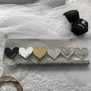 May include: A display of heart-shaped acrylic samples in black, white, gold, silver, frosted, and clear. Each heart is labeled with its color or finish. A gray rectangular tray holds the hearts, with a ring box and a ring visible in the background.