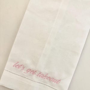 May include: White linen kitchen towel with pink embroidered text that reads "let's get takeout". The towel has a decorative stitched border along the bottom edge. The towel is folded and lies on a white surface.