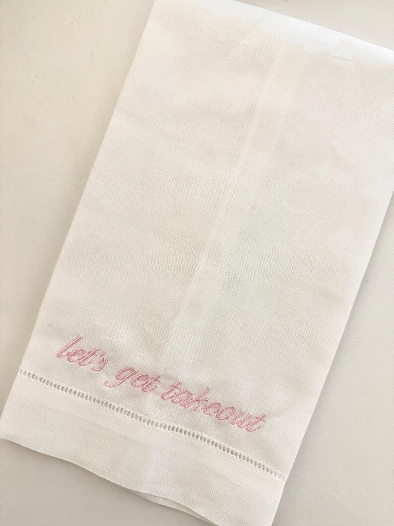 May include: White linen kitchen towel with pink embroidered text that reads "let's get takeout". The towel has a decorative stitched border along the bottom edge. The towel is folded and lies on a white surface.