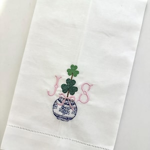 May include: White linen hand towel with a decorative embroidered design. The design features a blue and white vase with a shamrock and the letters "JS" in pink. The towel has a stitched border.