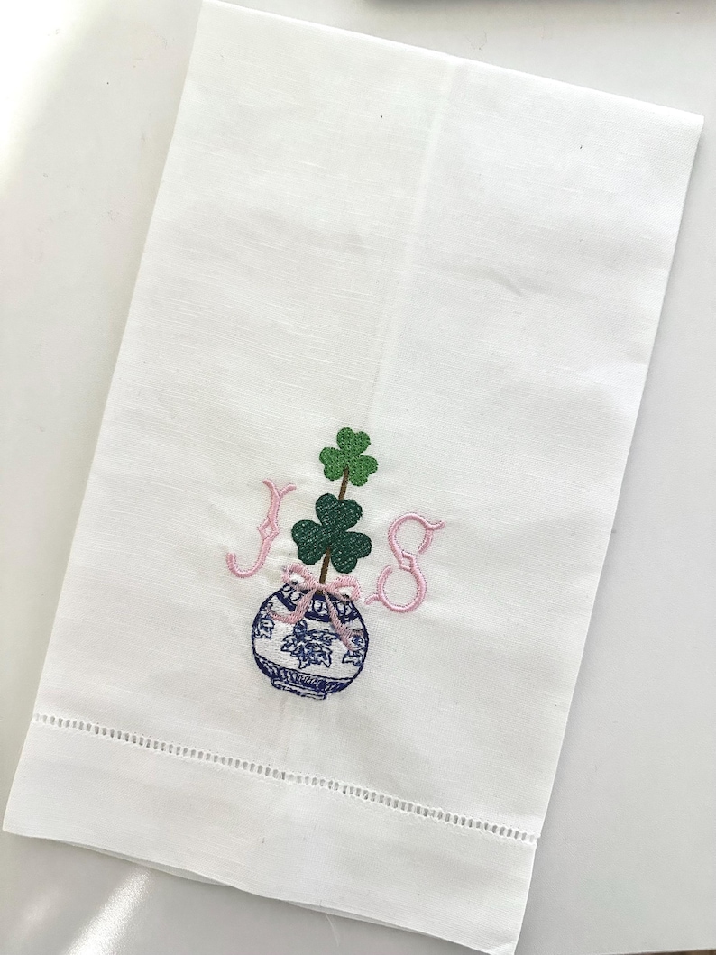 May include: White linen hand towel with a decorative embroidered design. The design features a blue and white vase with a shamrock and the letters "JS" in pink. The towel has a stitched border.