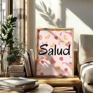 Puede incluir: Un cuadro enmarcado con la palabra "Salud" en escritura negra. El fondo es rosa con ilustraciones de rodajas de fruta y salpicaduras. El marco es de color madera clara, y el cuadro se exhibe sobre una mesa de madera.