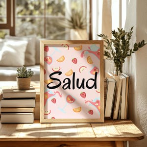 Puede incluir: Un cuadro enmarcado con la palabra "Salud" en escritura negra. El cuadro presenta un fondo rosa claro con ilustraciones de c&oacute;cteles, fresas y rodajas de naranja. El marco es de madera clara, y el cuadro est&aacute; sobre una mesa de madera.