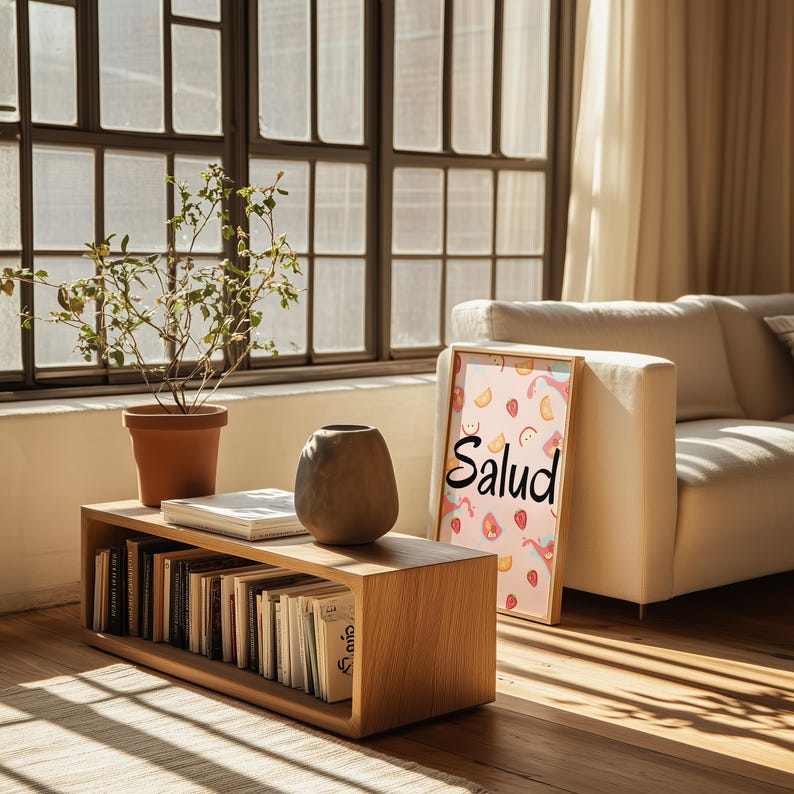 Puede incluir: Una escena de sal&oacute;n con una mesa de centro de madera que sostiene libros, un jarr&oacute;n y una planta en maceta. Una impresi&oacute;n enmarcada con la palabra "Salud" se apoya contra un sof&aacute; blanco. La luz del sol entra por una gran ventana.