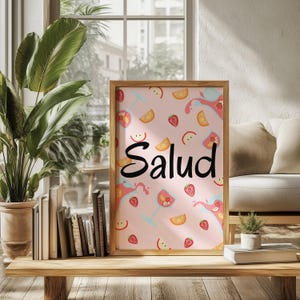 Puede incluir: Un estampado enmarcado con un marco de madera marr&oacute;n claro. El estampado tiene un fondo rosa con ilustraciones de frutas y c&oacute;cteles. La palabra "Salud" est&aacute; escrita en cursiva negra. El estampado se exhibe sobre una mesa de madera.