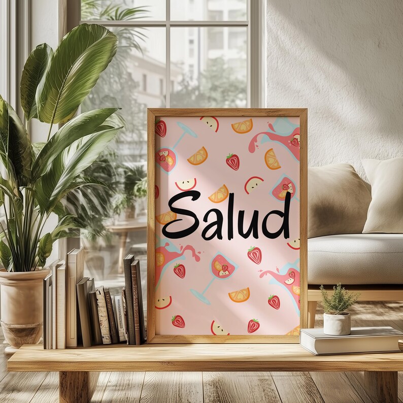 Puede incluir: Un estampado enmarcado con un marco de madera marr&oacute;n claro. El estampado tiene un fondo rosa con ilustraciones de frutas y c&oacute;cteles. La palabra "Salud" est&aacute; escrita en cursiva negra. El estampado se exhibe sobre una mesa de madera.