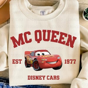 May include: Cream-coloured sweatshirt with the red text "MC QUEEN" and a red cartoon car graphic. The text "EST 1977 DISNEY CARS" is also printed on the shirt. The sweatshirt has a crew neck and long sleeves.