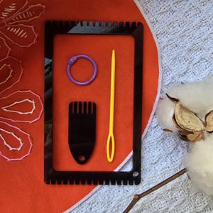 May include: A black plastic loom with a yellow needle, a purple ring, and a black comb-like tool. The loom is on a red fabric background.