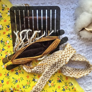 May include: A close-up of a loom with two strands of yarn, one white and one gold, being woven together. The loom is made of black plastic and has a grid of lines. A finished piece of woven fabric is lying on a yellow floral fabric.