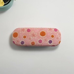 Puede incluir: Estuche rosa para gafas con un estampado floral y lunares coloridos en naranja, morado y rosa. El estuche tiene forma rectangular redondeada y está diseñado para proteger las gafas.