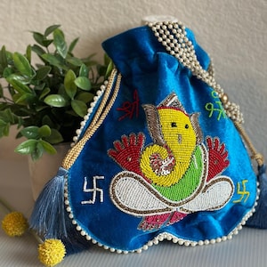 May include: A blue velvet pouch with an embroidered image of the Hindu deity Ganesha. The pouch features a beaded design with gold trim, tassels, and a pearl beaded drawstring. The image includes the Hindu symbol Swastika.