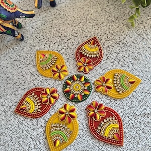 May include: A decorative rangoli set featuring a central circular piece surrounded by six teardrop-shaped pieces. Each piece is adorned with vibrant yellow, red, and green paint, intricate beadwork, and small clear crystals arranged in floral patterns.