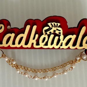 May include: A red wooden name badge with gold lettering that reads "Ladkewale". The badge has a gold chain with pearls and a gold design. The badge is likely for a wedding or special event.
